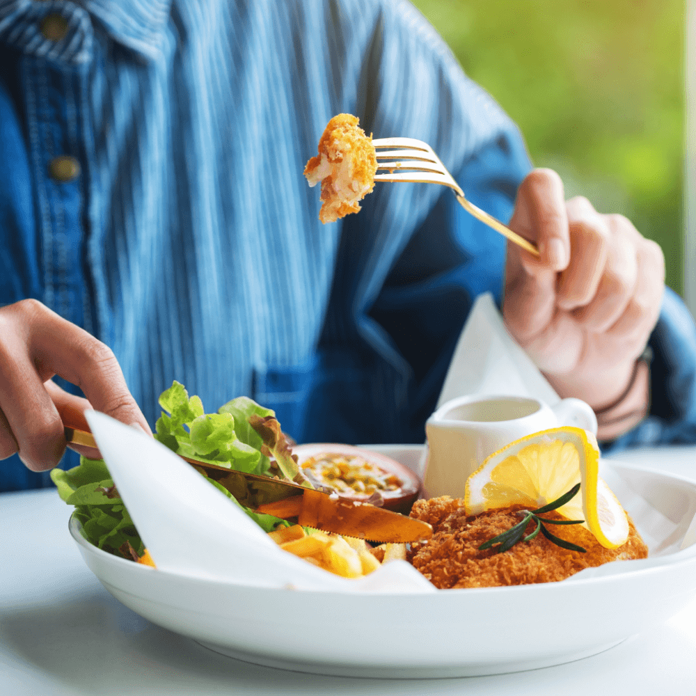 Person wearing a blue shirt eating breaded fried chicken with salad and lemon garnish, using a fork and knife. - Home Instead