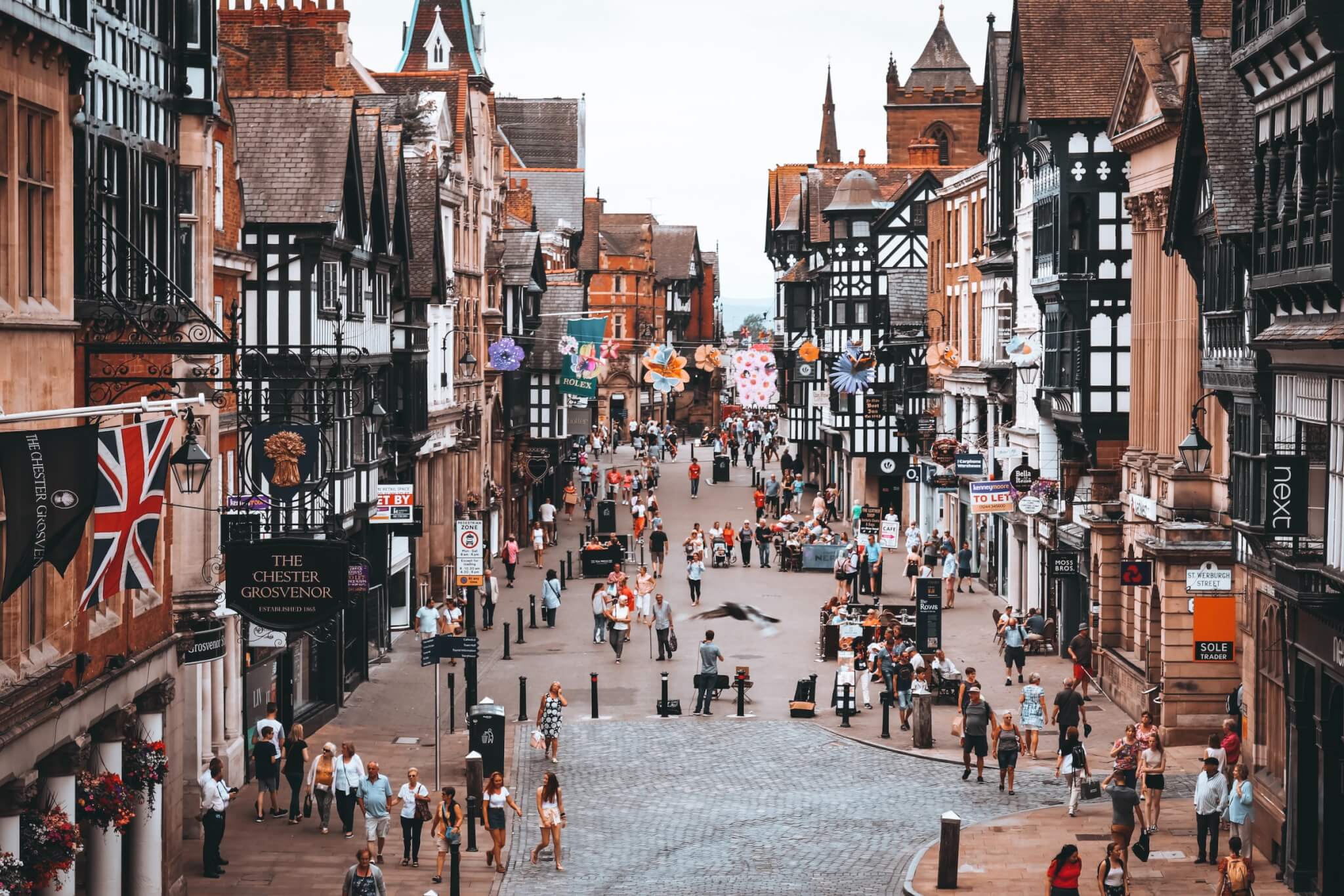 A bustling street scene in a historic town with old buildings, various shops, and numerous people walking around. - Home Instead