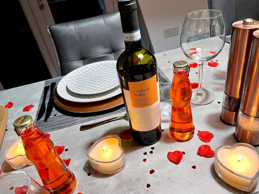 Romantic dinner setup with wine, soda bottles, lit candles, plates, and scattered heart-shaped confetti on the table. - Home Instead