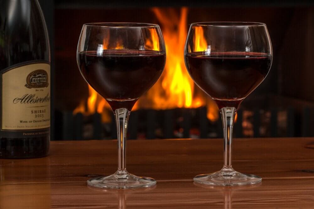 Two glasses of red wine on a wooden table in front of a roaring fireplace, with a wine bottle visible on the left. - Home Instead