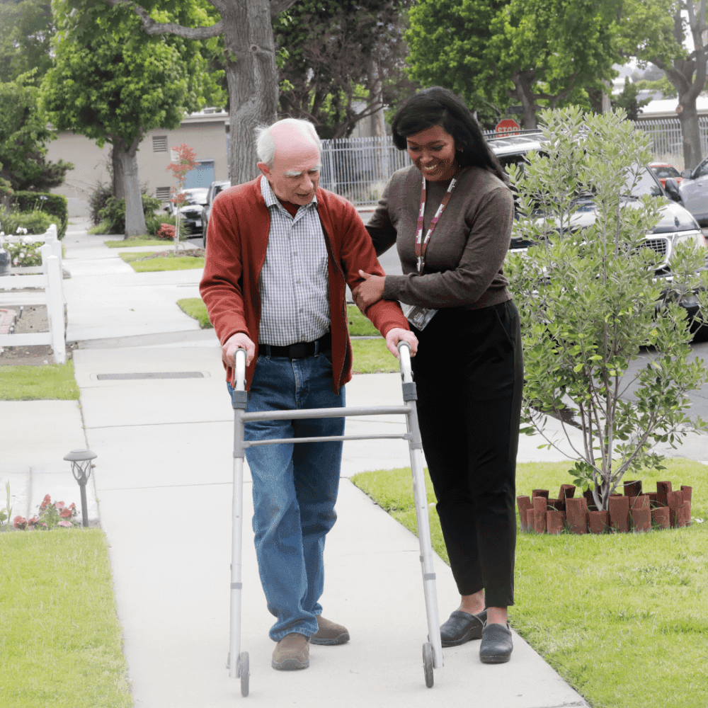 A caregiver assists a smiling elderly man using a walker outside on a sidewalk in a residential area. - Home Instead