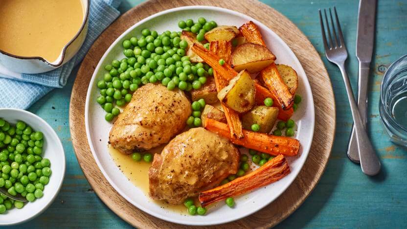 A meal with roasted chicken, peas, carrots, and potatoes on a plate with gravy in a bowl. Utensils are on the side. - Home Instead