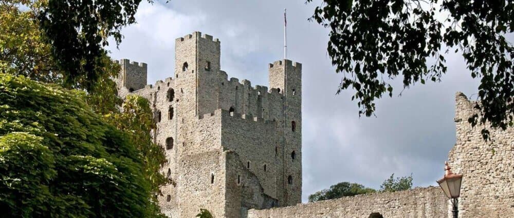 A medieval stone castle with tall turrets and an unfurled flag on a cloudy day, partially framed by leafy trees. - Home Instead