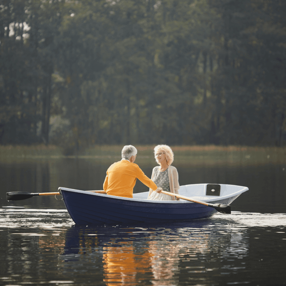 An elderly couple rowing a small blue boat on a calm lake surrounded by trees. - Home Instead