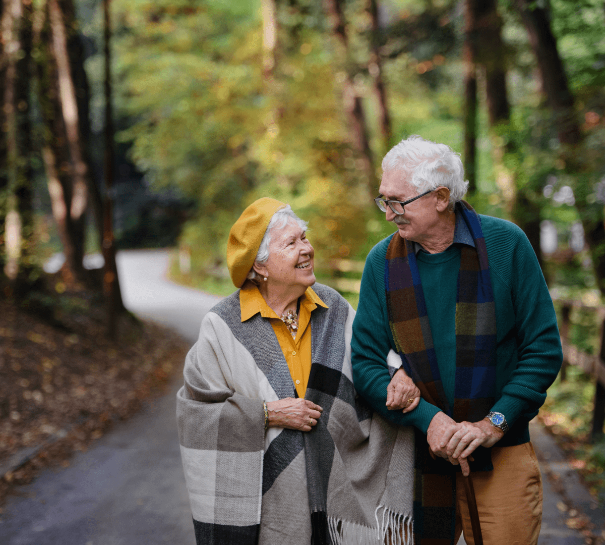 Elderly couple walking arm in arm on a forest path, wearing fall clothing, smiling and enjoying each other's company. - Home Instead