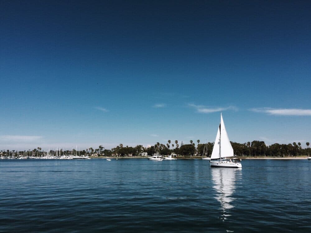 A sailboat glides on calm blue water with a distant shoreline of trees and boats under a clear blue sky. - Home Instead