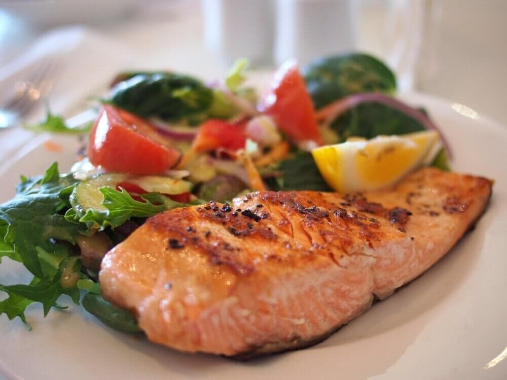Grilled salmon fillet served with a fresh salad of greens, tomatoes, red onion, and a lemon wedge on a white plate. - Home Instead