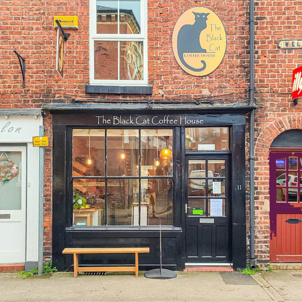 The front of a brick building with a sign reading "The Black Cat Coffee House," featuring a bench and a black door. - Home Instead