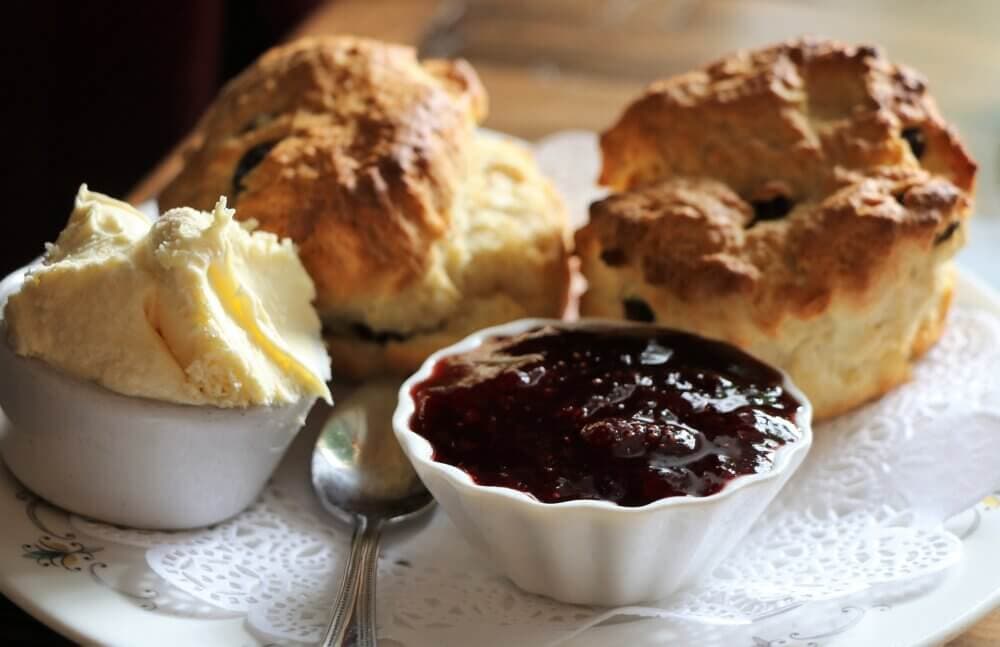 Two scones served with clotted cream and strawberry jam on a plate with a spoon. - Home Instead