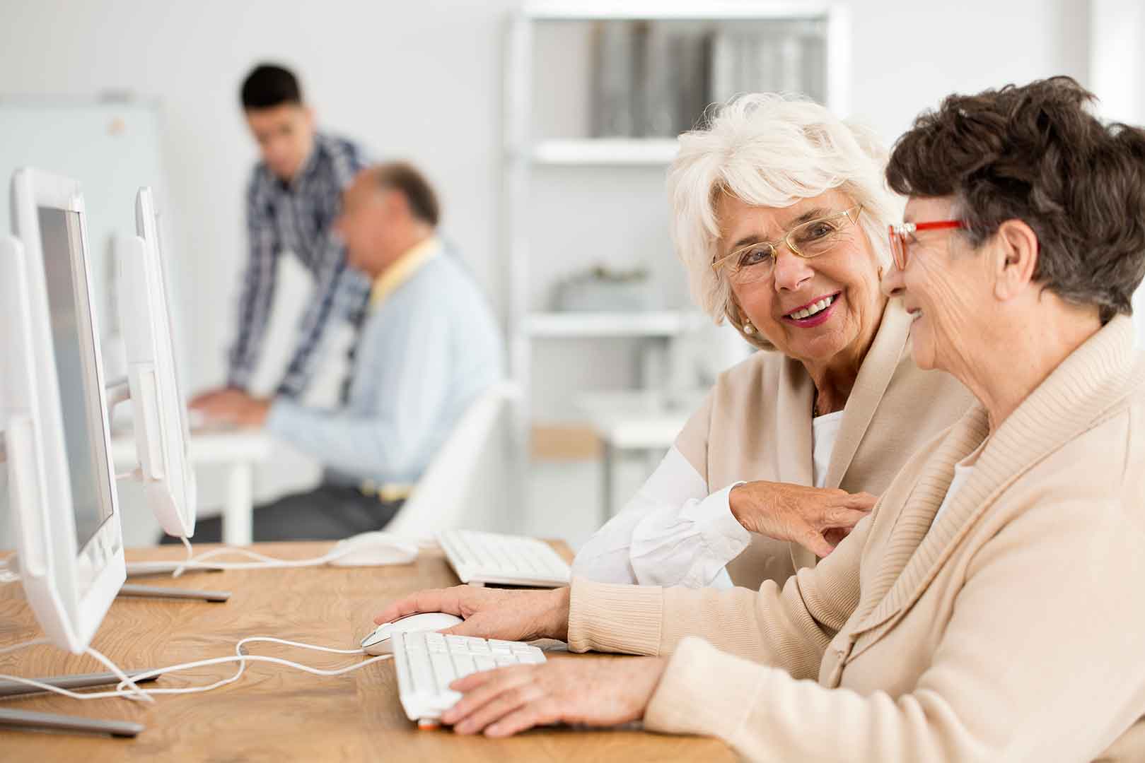 Two elderly women smiling and using computers in a classroom; two men working in the background. - Home Instead