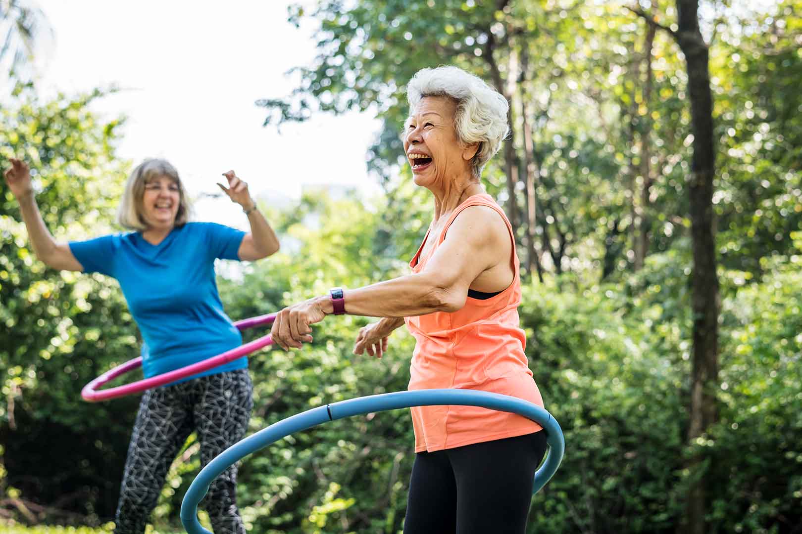 Two elderly women having fun hula hooping in a sunny, green outdoor park. They are smiling and laughing. - Home Instead