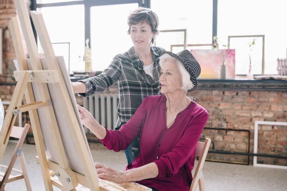 Older woman painting on canvas with younger woman assisting her in a well-lit art studio with brick walls. - Home Instead