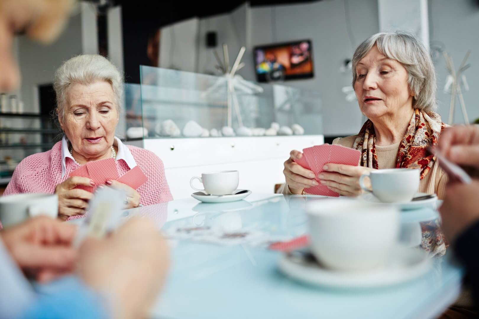 Two elderly women are sitting at a table, playing cards and drinking coffee in a bright, relaxed setting. - Home Instead