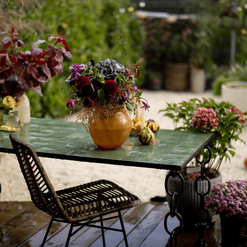 Flower arrangement in a vase sits on a green table outside, with a wicker chair beside it and plants in the background. - Home Instead