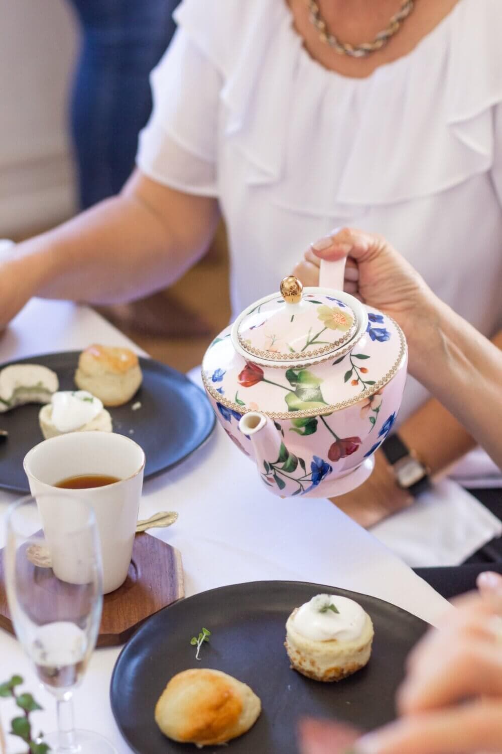 A person pours tea from a floral teapot at a table set with pastries, a cup of tea, and plates. - Home Instead