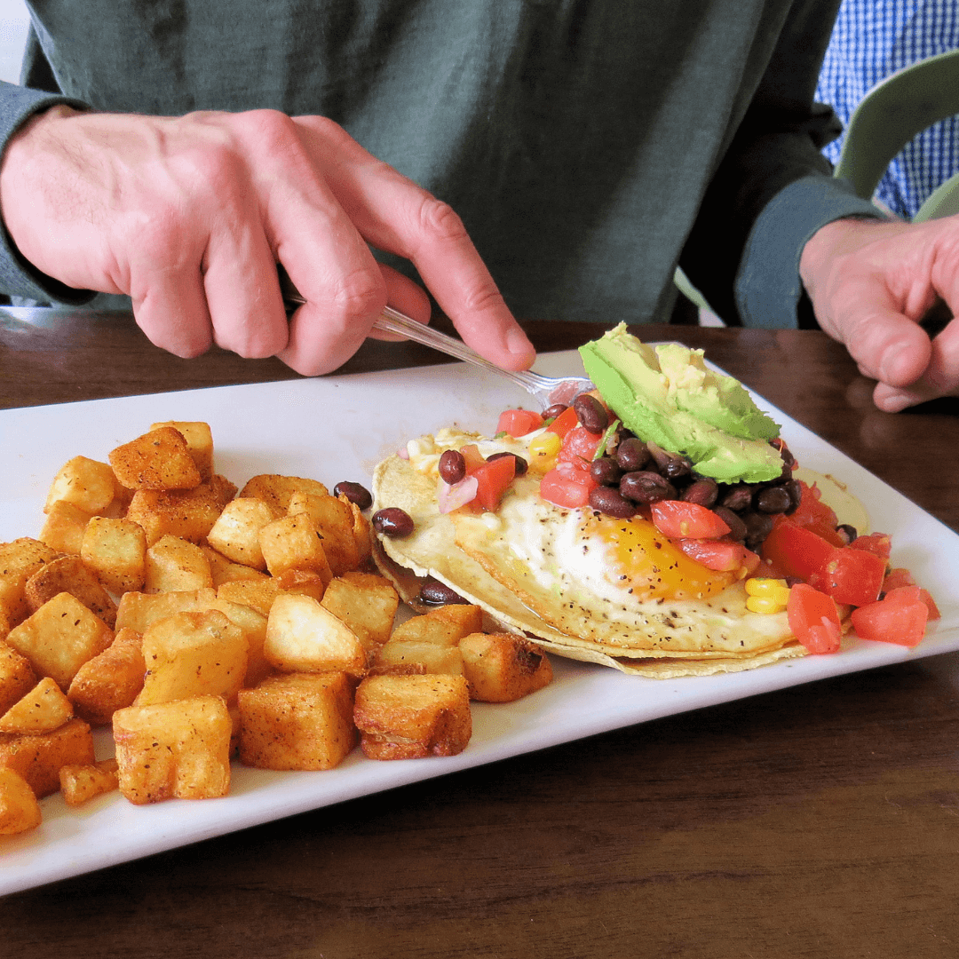 Person eating a dish with roasted potatoes, eggs, beans, avocado, and salsa on a white plate with a fork and knife. - Home Instead