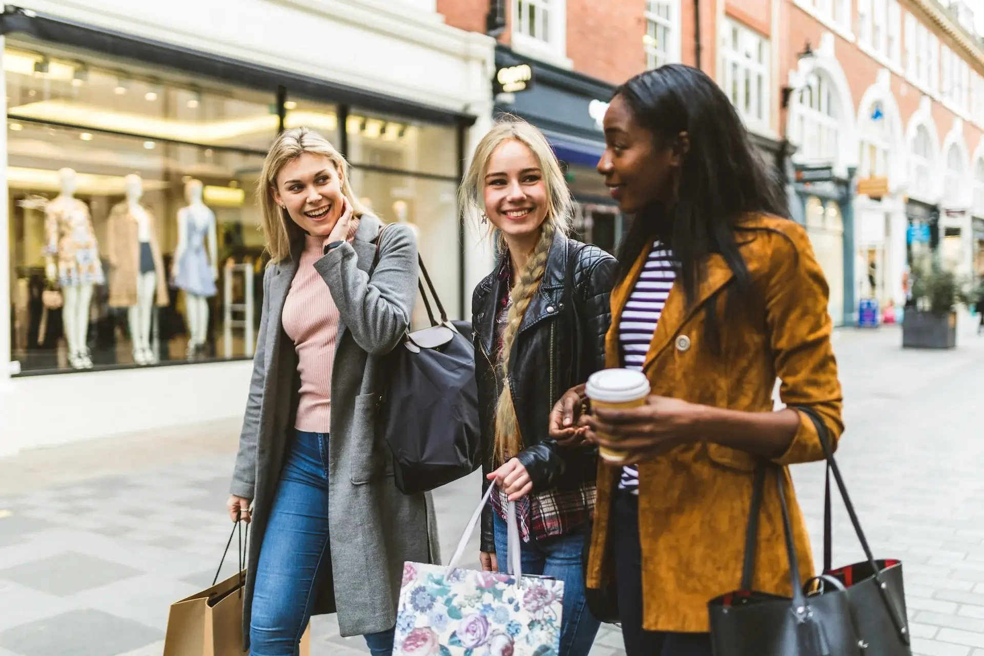 Three women walking and chatting happily on a shopping street, holding bags and a coffee cup. - Home Instead