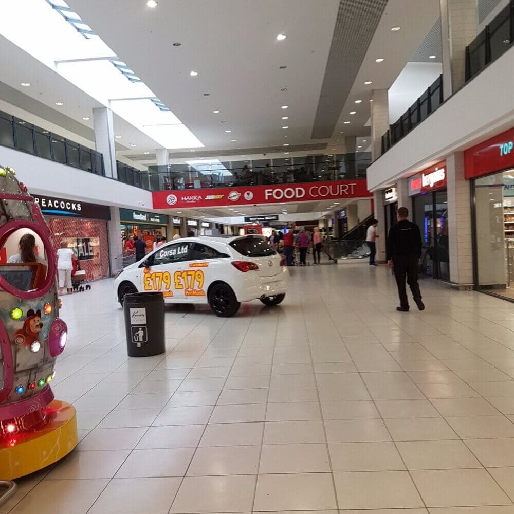 Interior of a shopping mall with a white car display, people walking, and a Food Court sign in the background. - Home Instead
