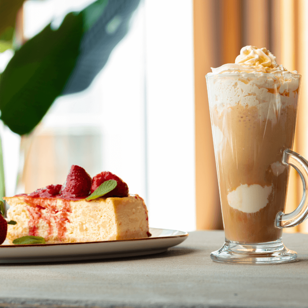 Slice of cheesecake with raspberries on a plate and a tall glass of iced coffee topped with whipped cream on a table. - Home Instead