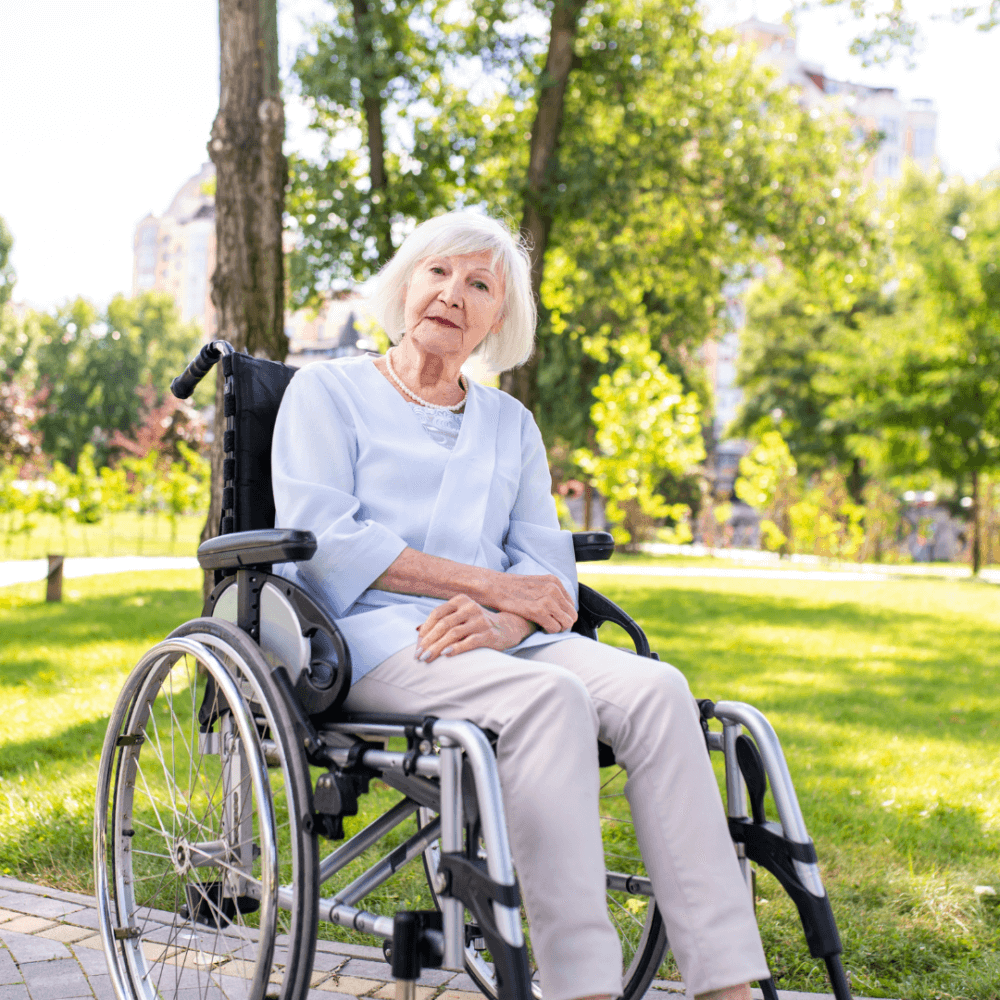 Elderly woman with white hair in a wheelchair, sitting in a park on a sunny day, wearing a light blue shirt and beige pants. - Home Instead