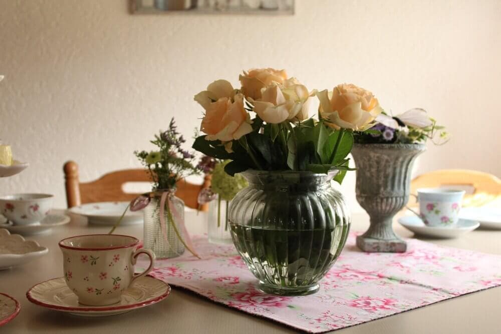 A dining table set with floral-themed tea cups, saucers, and a vase of light peach roses in the center. - Home Instead