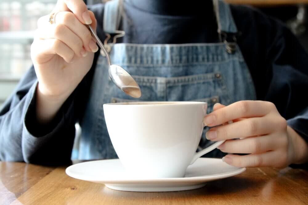 Person in blue overalls stirring a white cup of coffee with a spoon, at a wooden table. - Home Instead