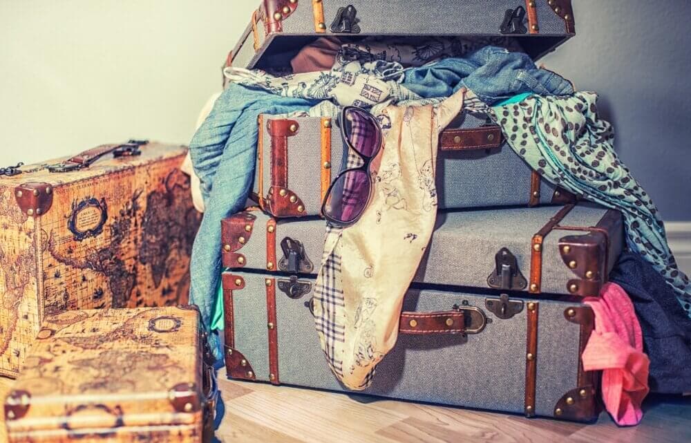 Stacked vintage suitcases with clothes and accessories spilling out, including scarves and sunglasses, placed on a wooden floor. - Home Instead