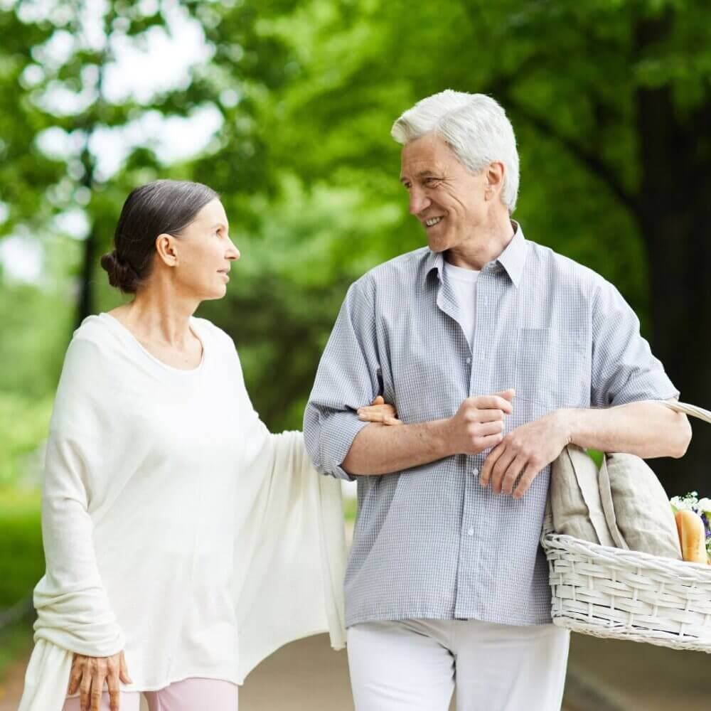 An elderly couple strolls in a park, arm in arm, wearing light-colored clothes, and carrying a basket. - Home Instead
