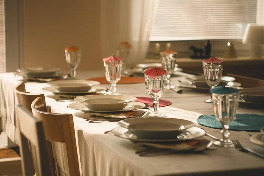 A dining table set for multiple people with plates, glasses with tiny umbrellas, and placemats in soft lighting. - Home Instead