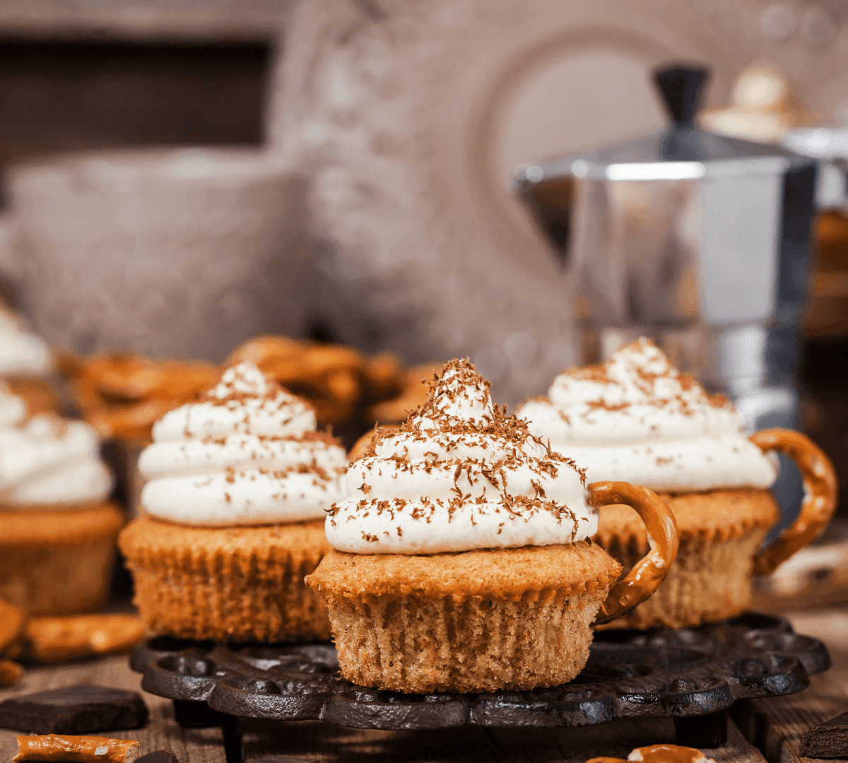 Cupcakes topped with whipped cream and chocolate shavings, decorated with pretzels. Coffee maker and dishes in background. - Home Instead