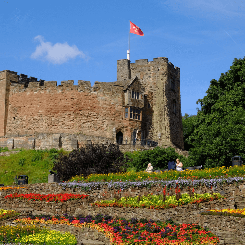 A historical castle with a red flag on top, surrounded by colorful flower gardens and lush green trees under a blue sky. - Home Instead