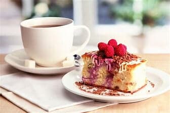 A cup of coffee and a slice of cake topped with raspberries and shaved chocolate on a table with a napkin. - Home Instead