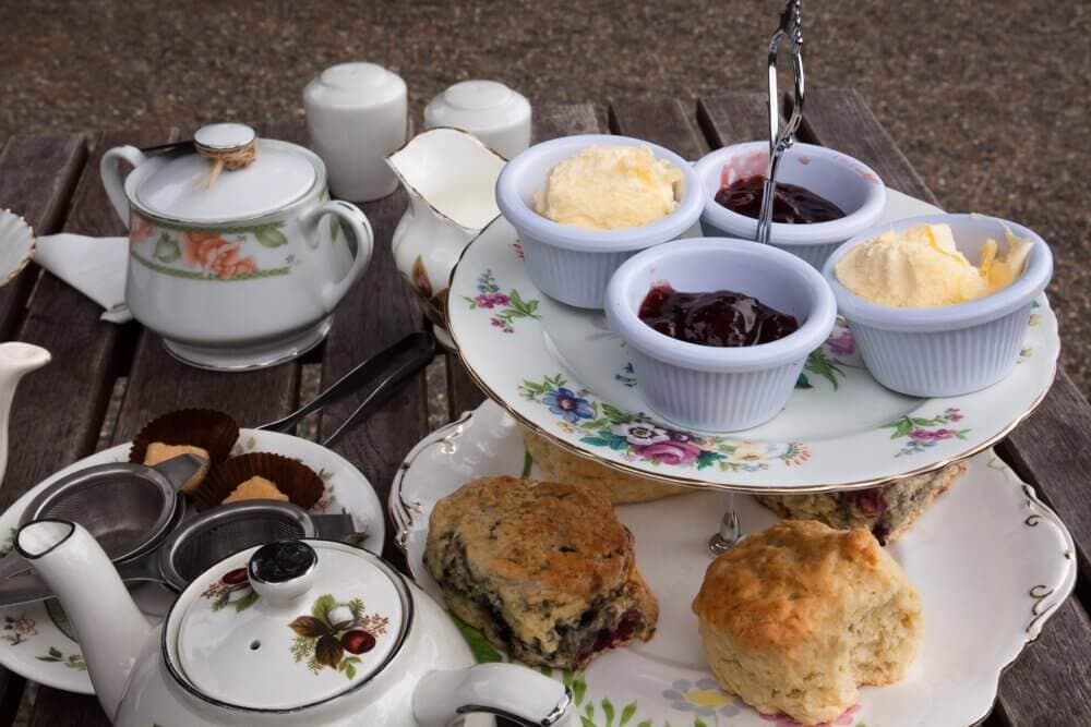A traditional English tea setup with scones, clotted cream, jam, teapots, and cups on a wooden table. - Home Instead