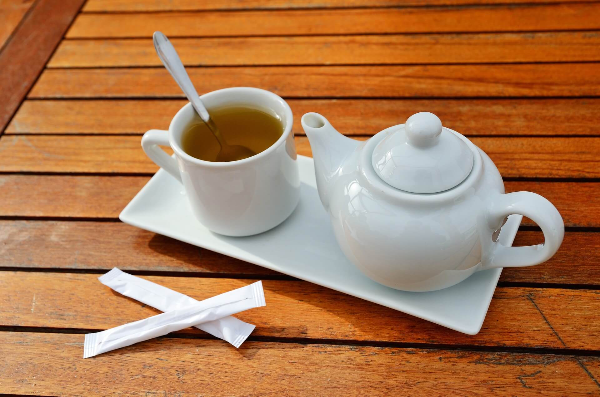 White teapot and cup with tea on a rectangular white tray, spoon in cup, and two sugar packets on wooden table. - Home Instead