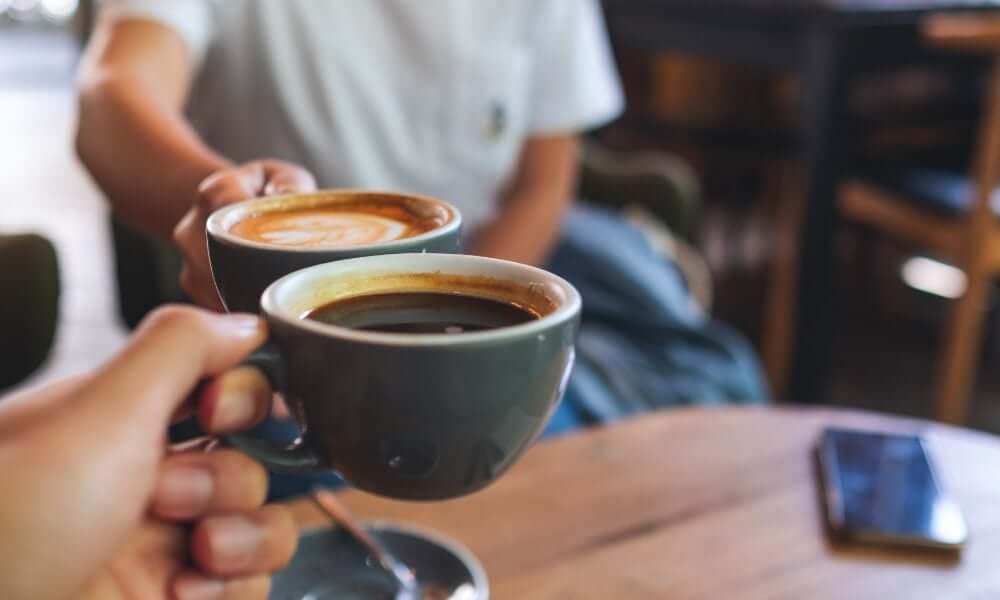 Two hands clinking coffee cups in a cozy cafe setting, with a blurred background and a smartphone on the wooden table. - Home Instead
