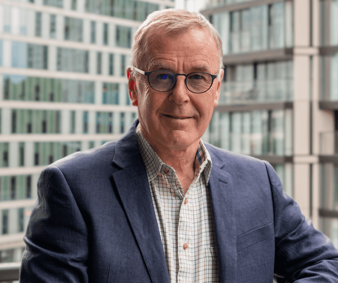 Elderly man with glasses in a blue blazer and checkered shirt, standing on a balcony with modern buildings in the background. - Home Instead