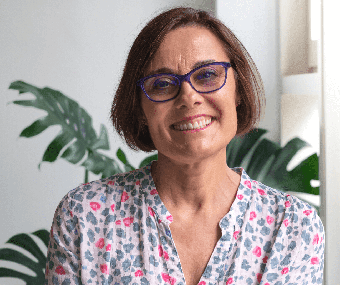 Smiling woman with short brown hair and blue glasses in a floral blouse, standing in front of a leafy plant. - Home Instead