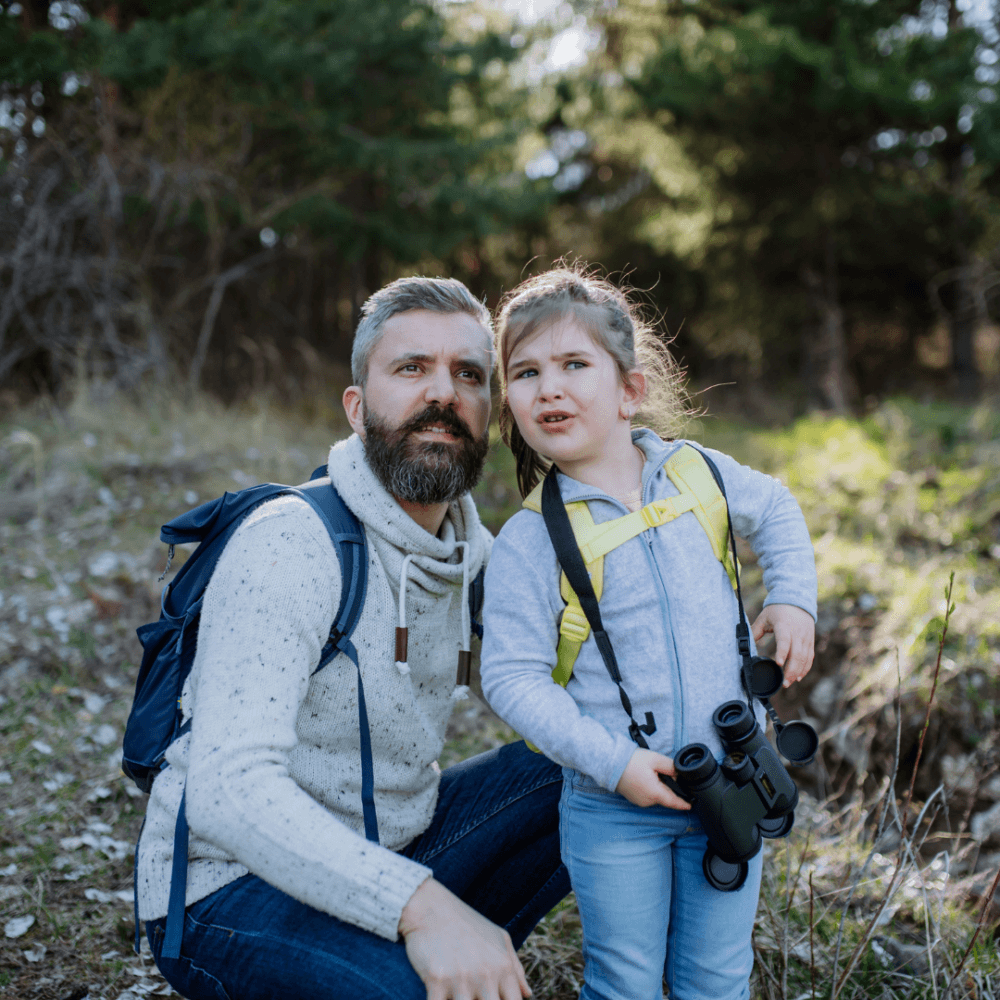 A bearded man with a young girl holding binoculars, both wearing backpacks, stand outdoors with trees in the background. - Home Instead