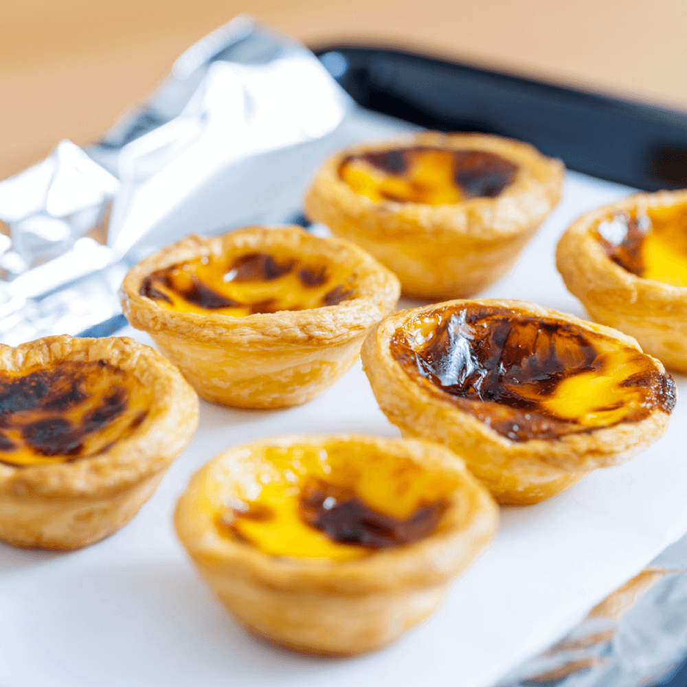 Close-up of six freshly baked Portuguese egg tarts on a tray lined with foil. - Home Instead