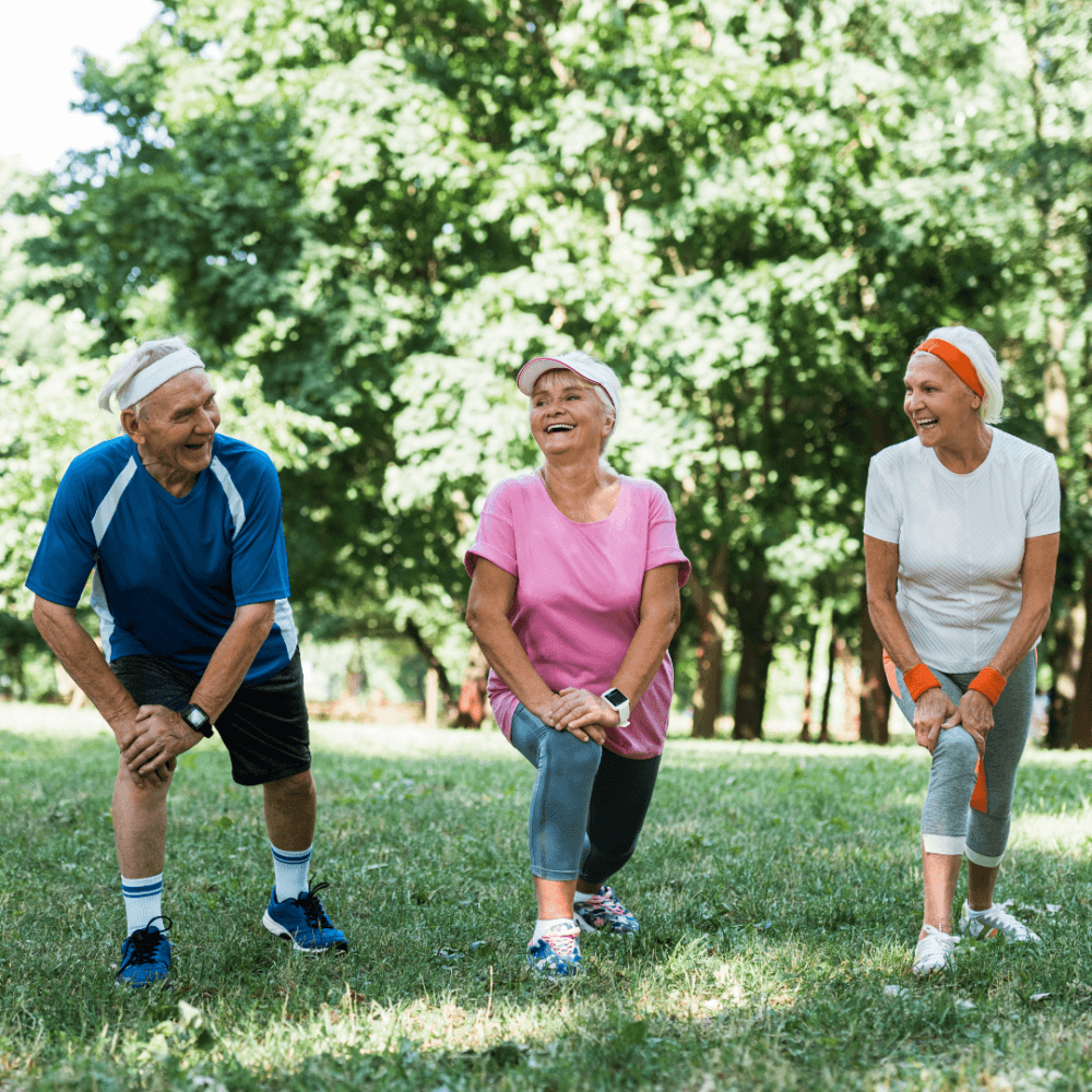 Three elderly individuals in sportswear laughing and stretching in a lush park. - Home Instead