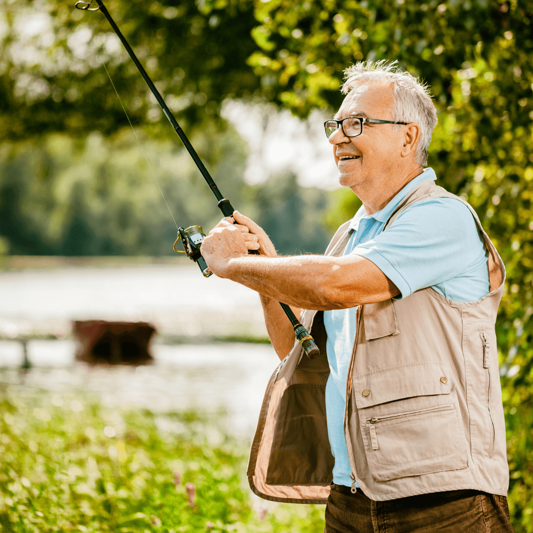 An elderly man in glasses and a fishing vest smiles while casting a fishing line into a lake on a sunny day. - Home Instead