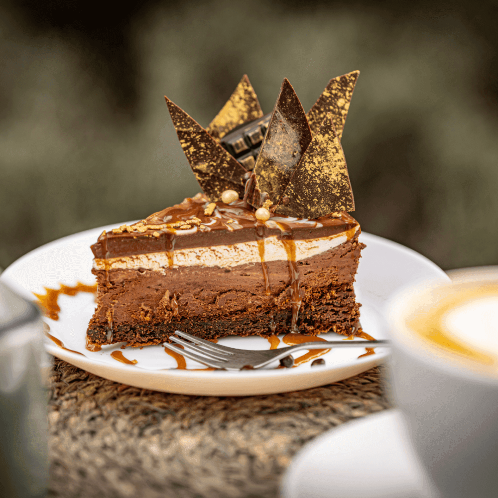 A slice of chocolate cheesecake topped with caramel drizzle and chocolate shards on a white plate with coffee in the foreground. - Home Instead