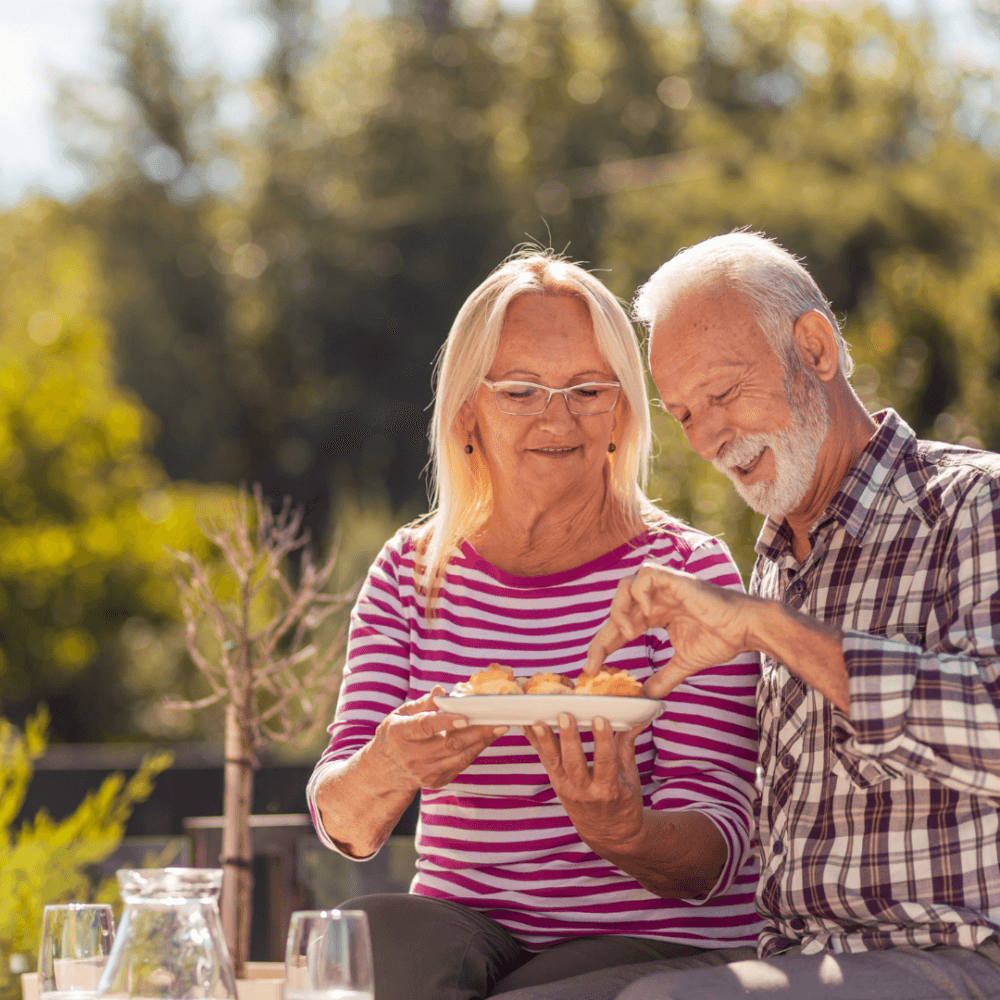 A senior couple enjoys a meal outdoors, smiling and sharing food under bright daylight with lush greenery around them. - Home Instead