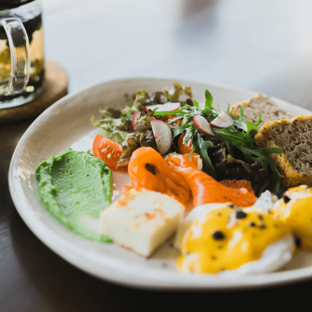 A plate with avocado, smoked salmon, feta, poached eggs, salad, and bread, with a drink in the background. - Home Instead