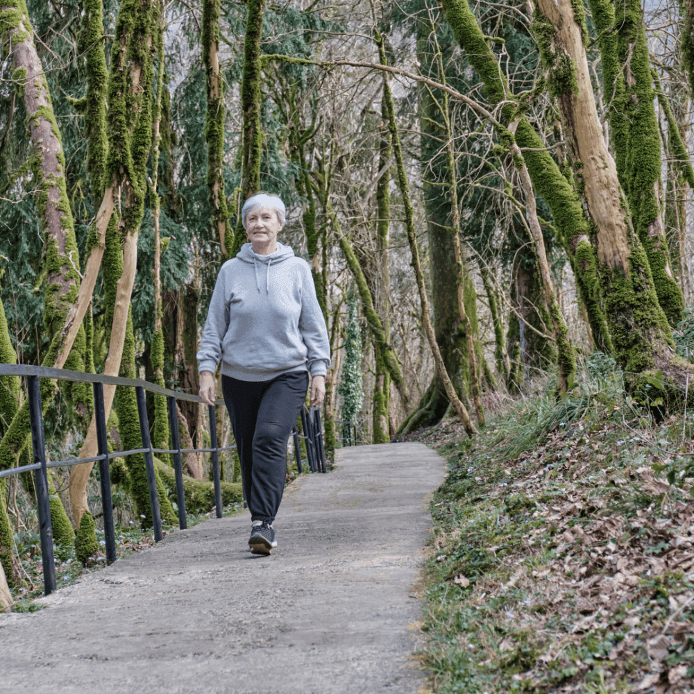 Elderly woman with short hair walks along a forest path with one hand in her pocket, surrounded by trees and moss. - Home Instead