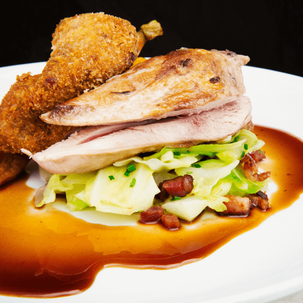 A gourmet dish featuring roasted poultry, sautéed cabbage with pancetta, and two breaded and fried croquettes, garnished with sauce. - Home Instead