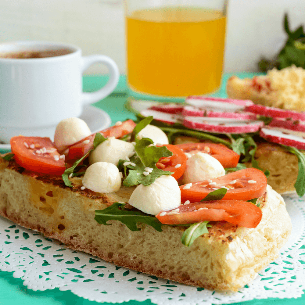 Slice of bread with tomatoes, mozzarella, and arugula; radish, orange juice, and coffee in the background. - Home Instead