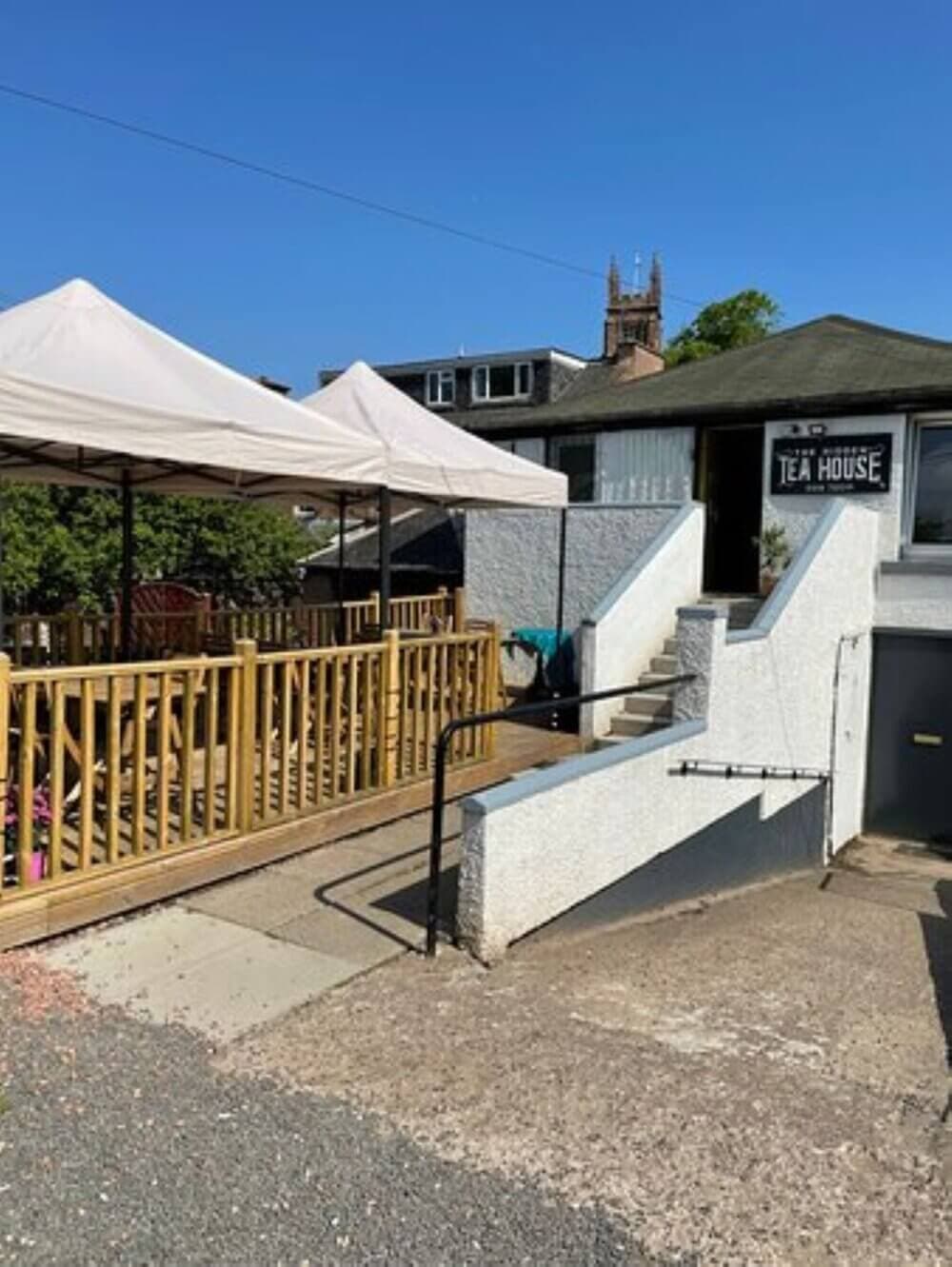 Outdoor seating area of a teahouse with a gazebo-covered deck, ramp, and stairs, with a church tower in the background. - Home Instead