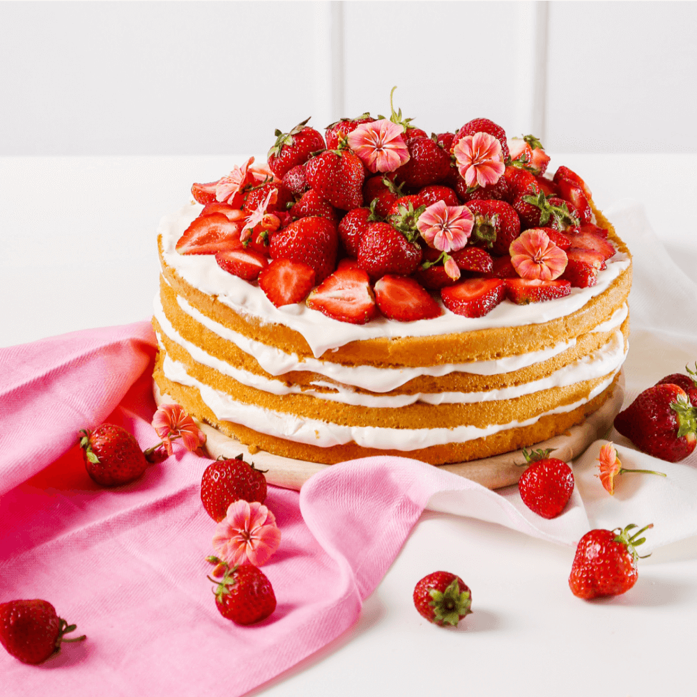 Four-layer vanilla cake with cream, topped with fresh strawberries and pink flowers, and surrounded by a pink cloth. - Home Instead