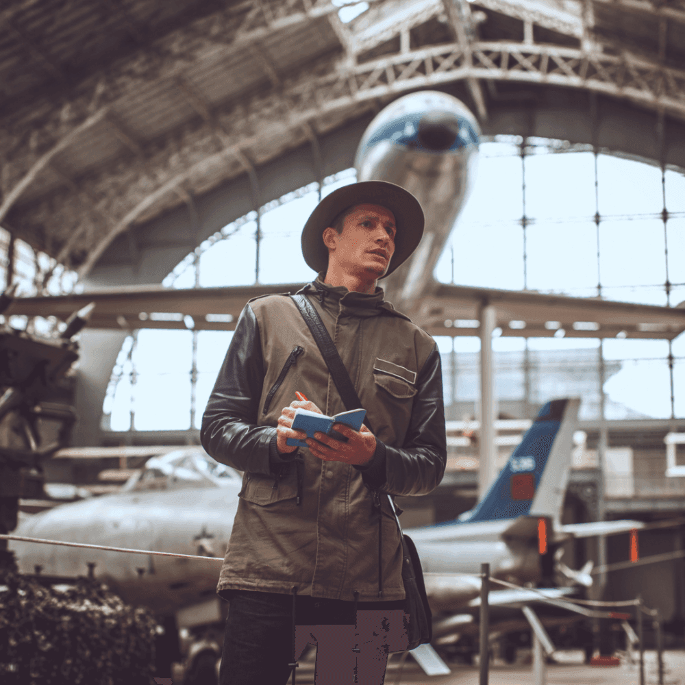 A man in a hat takes notes in an aviation museum with aircraft in the background. - Home Instead